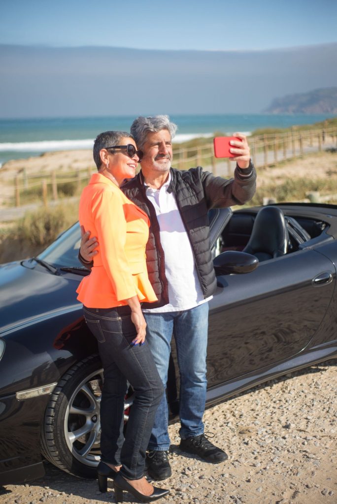 A happy senior couple taking a selfie by a car on a scenic coastal road in Portugal.